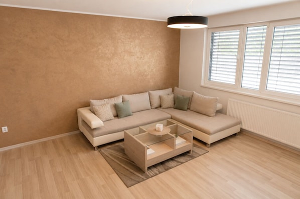 A modern living room with a sectional sofa, a glass coffee table, and a large window with blinds.