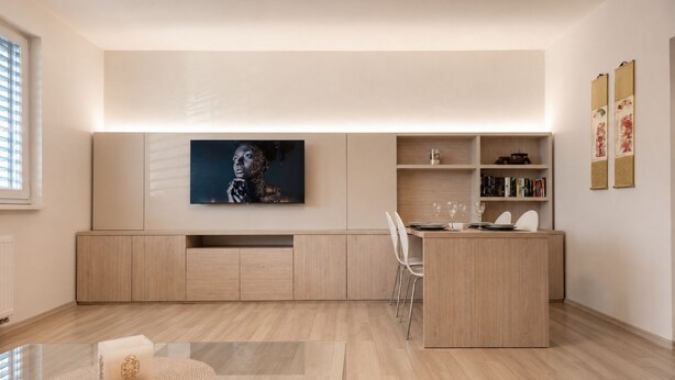 A modern kitchen with a dining area, wooden cabinetry, a TV mounted above the counter, and a bookshelf with books. - Apartment Pred Polom (Trenčín)