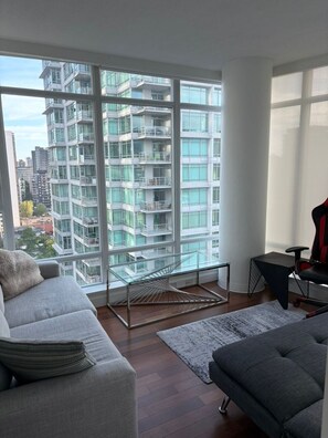 A modern living room with a large window, a glass table, a sofa, and a gaming chair. - Coalharbour Vantage Point 1616 (Vancouver)