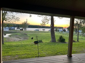 Une véranda couverte offrant une vue sur une cour verdoyante, un étang et des maisons situées de l’autre côté d’une étendue d’eau, au coucher du soleil.