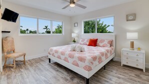 Une chambre à coucher comprenant un lit, une chaise, une table de chevet, un téléviseur et des fenêtres donnant sur des palmiers.