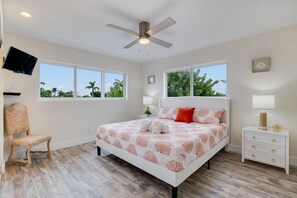 A bedroom with a bed, a chair, a nightstand, a TV, and windows with a view of palm trees. - Sanibel Arms A8- Gorgeous Condo Steps to Beach (Sanibel)