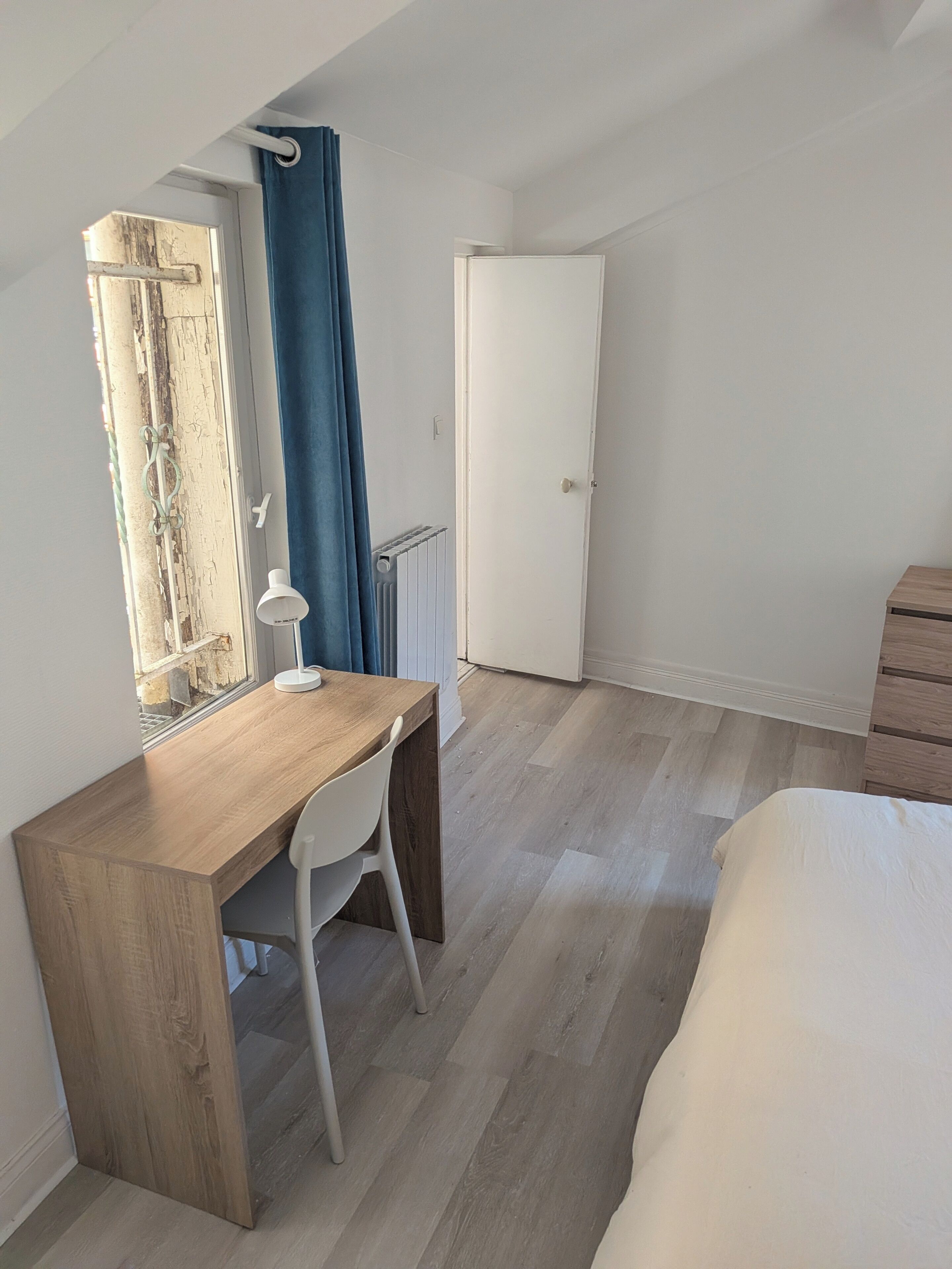 Ein Schlafzimmer mit einem Holztisch, einem weißen Stuhl, einem Bett, einer weißen Tür und einem Fenster mit Vorhang.