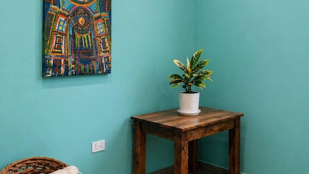 Una habitación con paredes color turquesa, una mesa de madera con una planta en maceta, un cuadro abstracto colorido, una silla de mimbre con una manta y un equipo de aire acondicionado.