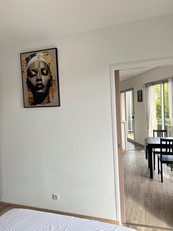 Una habitación con pared blanca, una obra de arte enmarcada, suelo de madera, una cama y un umbral que da a otra habitación con una mesa y sillas.