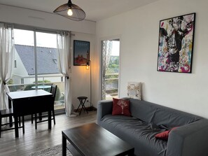 Una sala moderna con un sofá gris, una mesa de centro de madera, una mesa de comedor con sillas, una mesita auxiliar pequeña y un ventanal grande con cortinas.