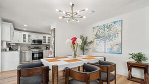 Eine moderne Küche mit Edelstahlgeräten, eine Essecke mit weißem Tisch und Holzstühlen sowie eine Deckenleuchte.