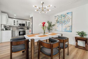 Eine moderne Küche mit Edelstahlgeräten, eine Essecke mit weißem Tisch und Holzstühlen sowie eine Deckenleuchte.