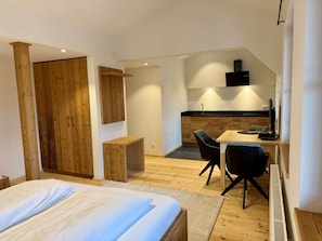 Ein Schlafzimmer mit Bett, einem Holzschrank, einem kleinen Tisch, einem Stuhl und einer Küchenzeile mit Spüle und Herd.