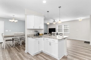 A modern kitchen with white cabinetry, a dining area with a wooden table and chairs, and pendant lighting. - "Coastal Belle" By Beach Star Near Marsh Walk (Murrells Inlet)
