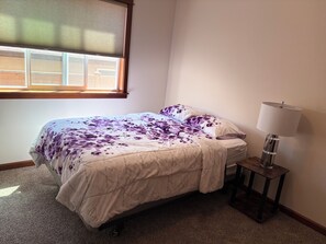 Ein Schlafzimmer mit einem Bett, einem Fenster mit Jalousien, einer Nachttischlampe und Teppichboden.