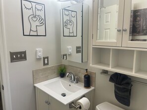 Un baño con lavamanos blanco, espejo y retrete. Hay interruptores de luz en la pared y un dispensador de jabón.