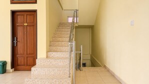 Una escalera con barandilla, una puerta de madera con placa de número y un cubo de basura verde.