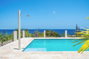 A rectangular pool with a clear blue sky and ocean view, surrounded by a fence and vegetation. - Vrbo Property (Kralendijk)