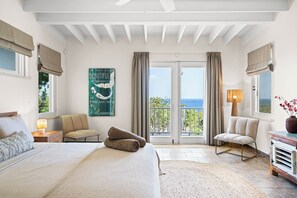 Een slaapkamer met een bed, twee stoelen, een nachtkastje, een lamp, een schilderij en uitzicht op zee.