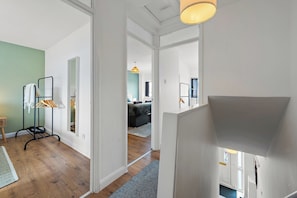 A hallway with a rack of clothes, a staircase, and a living room in the background. - Cosy 3-Bed House Basildon | Contractors | Parking (Vange)