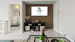 Un salon moderne avec un téléviseur fixé au mur qui affiche le logo d’une société de gestion immobilière, un meuble de rangement noir pour appareils multimédias et une table basse en verre sur laquelle sont posés des verres et des bouteilles de vin.