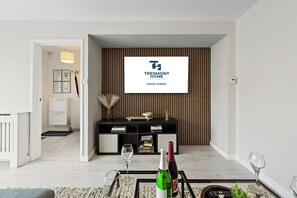 A modern living room with a mounted TV displaying a property management company, a black entertainment unit, and a glass coffee table with wine glasses and bottles. - Cosy 3-Bed House Basildon | Contractors | Parking (Vange)