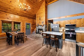 A wooden cabin interior with a dining area, kitchen, and fireplace. - NEW! Hidden Cabin with Mountain Views, Hot Tub, Fire Pit, Gameroom & More (Maggie Valley)