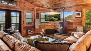 Ein rustikales Blockhaus-Interieur mit Kamin, Holzwänden, einer Couch und einem großen Fenster mit Aussicht.