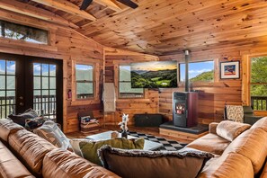 A log cabin interior with a fireplace, wooden walls, a sofa, and a large window with a view. - NEW! Hidden Cabin with Mountain Views, Hot Tub, Fire Pit, Gameroom & More (Maggie Valley)