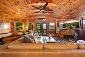 A log cabin interior with a leather sofa, a fireplace, and a large window with a scenic view. - NEW! Hidden Cabin with Mountain Views, Hot Tub, Fire Pit, Gameroom & More (Maggie Valley)