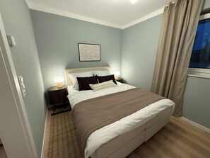 Un dormitorio con cama, lámparas de noche, un tapete, un cuadro enmarcado en la pared y una ventana con cortinas.