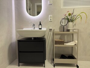 Un baño con lavamanos blanco, tocador negro, espejo, planta en maceta y repisa con toalla y cesta.