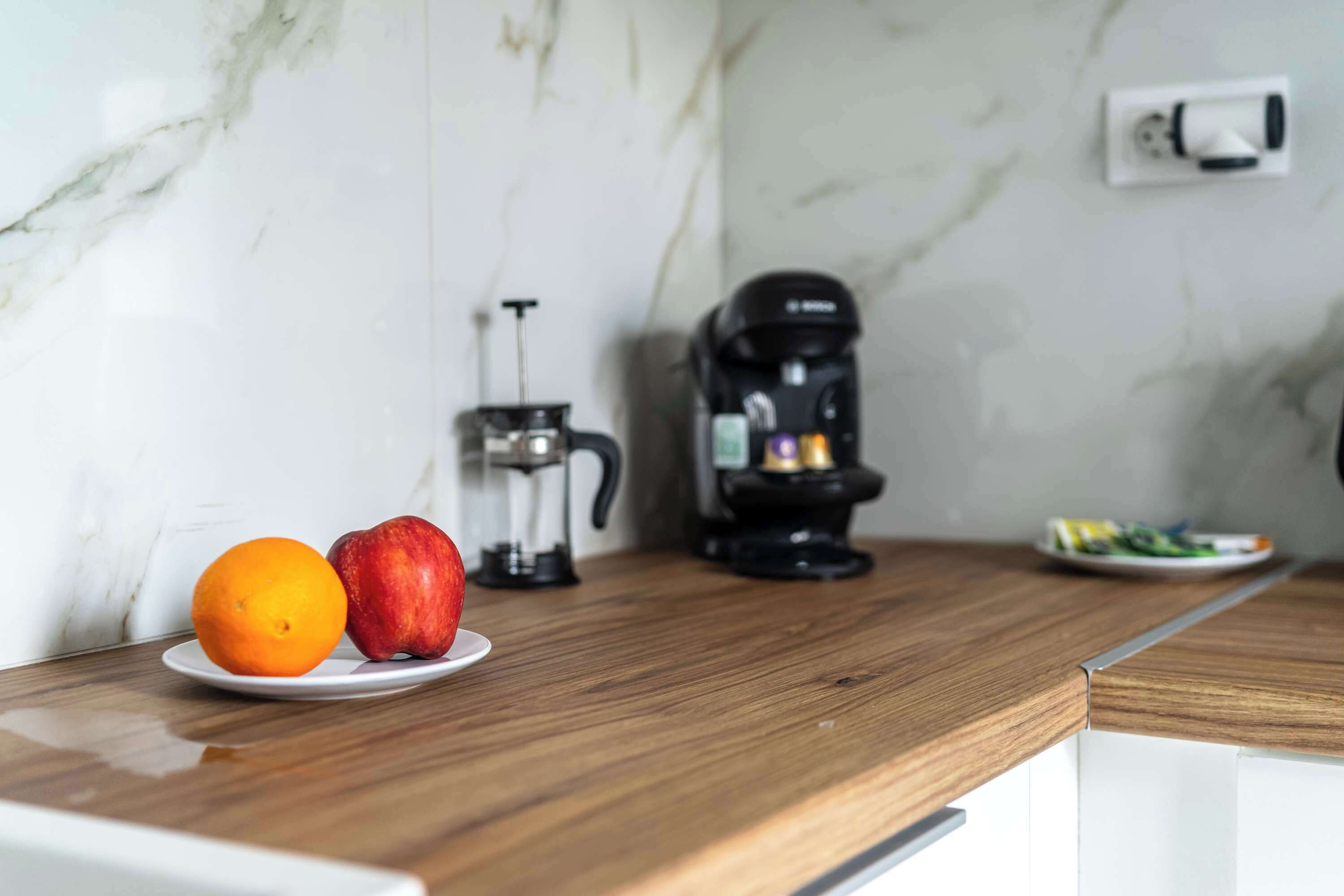 A kitchen counter with a coffee maker, a plate with an orange and an apple, and a coffee pot.