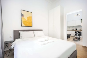 Ein modernes Hotelzimmer mit einem ordentlich bezogenen Bett, einem kleinen Nachttisch, einem an der Wand hängenden Kunstwerk und Blick in den Küchenbereich.