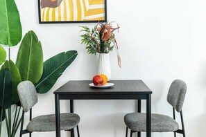 Uma mesa de jantar com cadeiras, um vaso com plantas e frutas, e um quadro na parede.