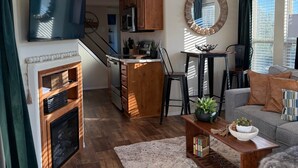 A spacious loft-style living area with a high ceiling, wooden flooring, a fireplace, a kitchen with wooden cabinets, a dining area, and a balcony with a railing.