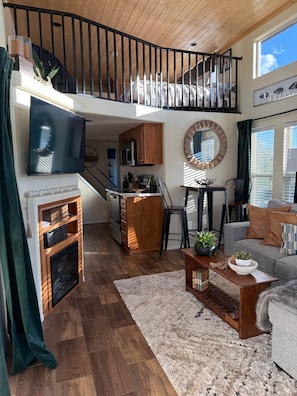 Amplio espacio tipo loft con techo alto, piso de madera, chimenea, cocina con gabinetes de madera, comedor y balcón con barandilla.