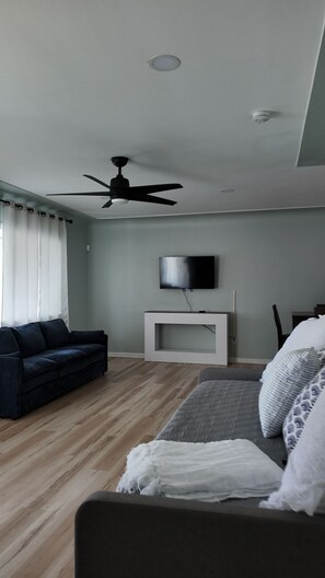 A modern living room with a ceiling fan, a flat-screen TV mounted on a white console, a dark blue sofa, and a grey sofa with patterned cushions. - House in Seminole with AC, close to beach, private pool, fenced yard for privacy (Seminole)