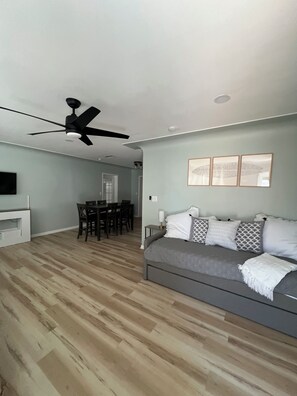Una sala moderna con un sofá gris, un ventilador de techo, una zona de comedor con sillas y un televisor sobre un mueble blanco.