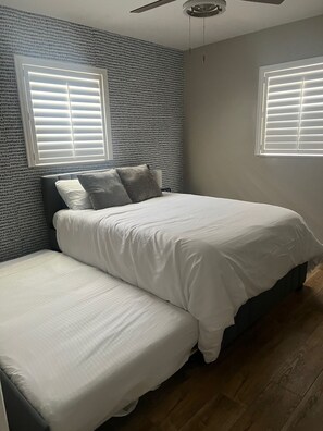 Un dormitorio con cama, dos almohadas, ventilador de techo y ventana con persianas.