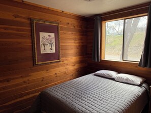 Quarto com paredes de madeira, cama com colchão acolchoado, ilustração botânica emoldurada e janela com cortinas.