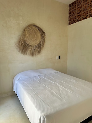 Una cama individual con colchón blanco y una manta con textura, un tapiz de pared tejido, y un enchufe en la pared.