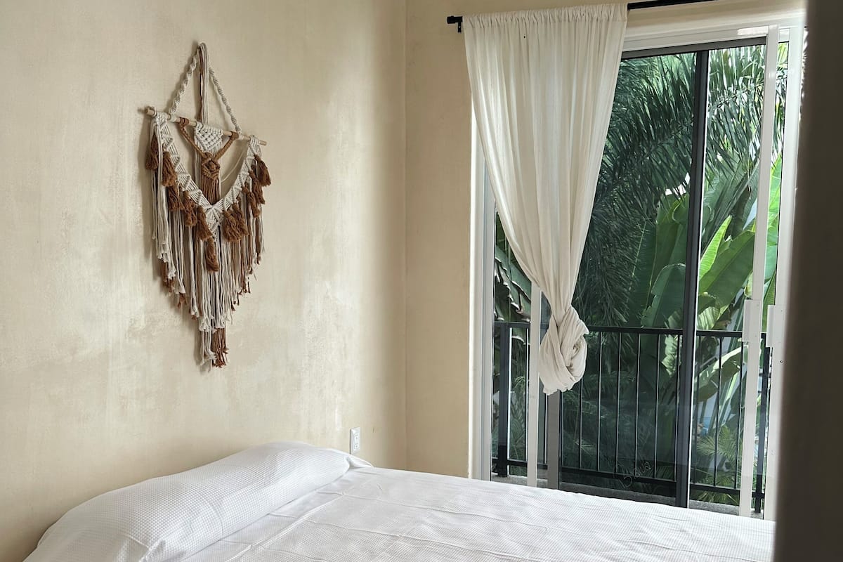 A bedroom with a bed, a macrame wall hanging, and a window with curtains.
