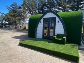 A small, white, arched structure with a green roof, a door, and two small green benches in front.
