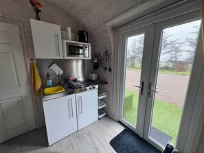 A compact kitchen with white cabinets, a microwave, and a sink. There is a door leading to a balcony with a view of a grassy area. - Sea Thrift, Coastal Pod, near Oldshoremore Beach (Lairg)