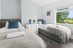 A bedroom with two beds, a nightstand, a window with a view, and patterned wallpaper. - Spacious Home with Garden for Groups & Families (Greater Manchester)