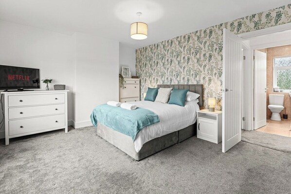 A bedroom with a bed, bedside tables, a TV, and a bathroom visible through an open door. - Spacious Home with Garden for Groups & Families (Greater Manchester)