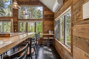 A wooden interior with a dining table, chairs, and large windows offering a view of the forest.