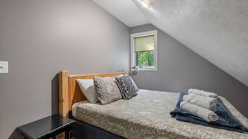 Une chambre avec un plafond en pente, un lit recouvert d’une couvre-lit à motifs, deux oreillers et une fenêtre donnant sur un paysage verdoyant.