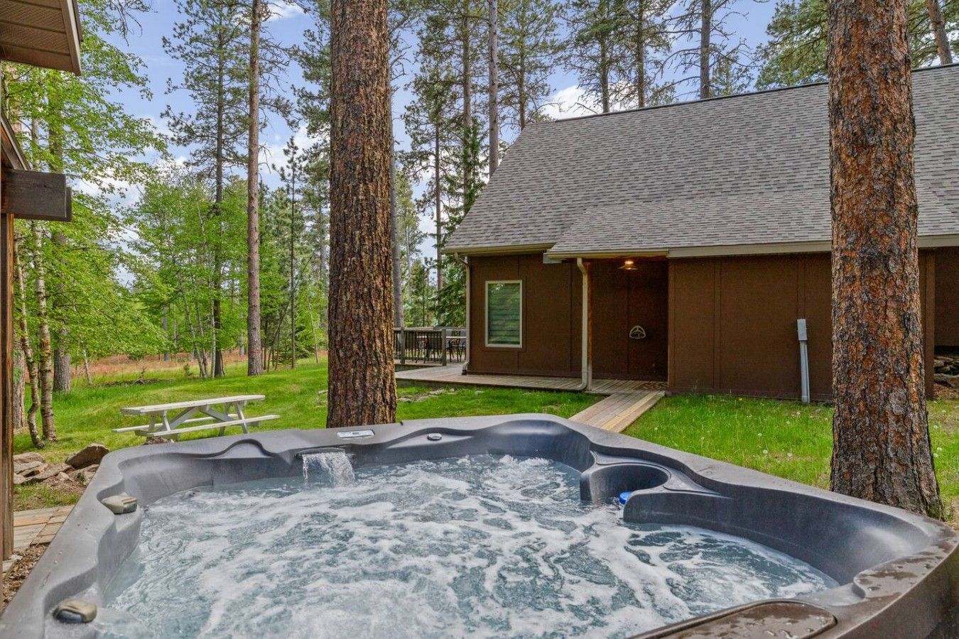 A hot tub surrounded by trees, a wooden deck, and a cabin with a porch.