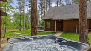 A hot tub surrounded by trees, a wooden deck, and a cabin with a porch.
