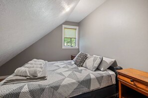 A bedroom with a sloped ceiling, a bed with a geometric-patterned bedspread, a window with a view of trees, and a wooden nightstand.