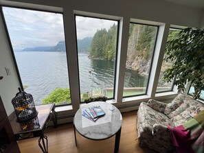 Ein Zimmer mit großen Fenstern, das einen Blick auf einen See und Berge bietet, ein runder Tisch mit Zeitschriften, ein blumiges Sofa und ein schwarzer Metallstuhl.