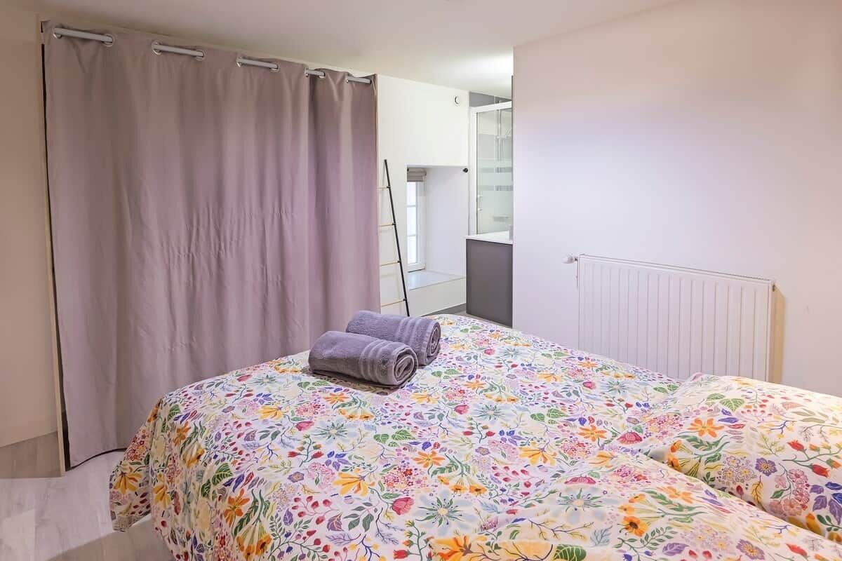 Une chambre à coucher avec un lit, des rideaux, un radiateur, et une salle de bain visible au fond.
